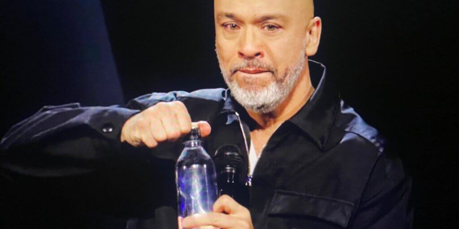 Filipino comedian Jo Koy is shown opening a water bottle and holding a microphone. His lips are pursed, his eyes are misty with tears.