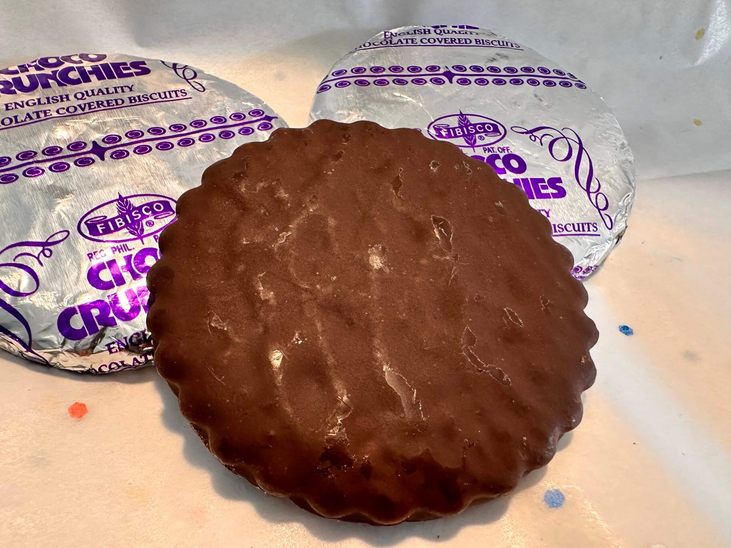 Two foil-wrapped, flat chocolates with wrappers saying Choco Crunchies, with one unwrapped, scalloped edge, flat chocolate.